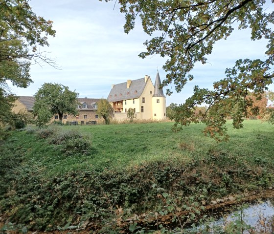 Burg Langendorf umgeben von gr&uuml;ner Wiese und B&auml;umen, mit einem kleinen Bach im Vordergrund. Der Himmel ist leicht bew&ouml;lkt., &copy; Paul Meixner