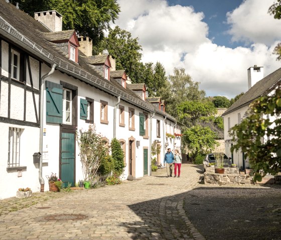Gepflasterte Stra&szlig;e mit historischen Fachwerkh&auml;usern in Kronenburg. Zwei Personen spazieren, umgeben von bl&uuml;henden Pflanzen und B&auml;umen., &copy; Eifel Tourismus GmbH, Dominik Ketz