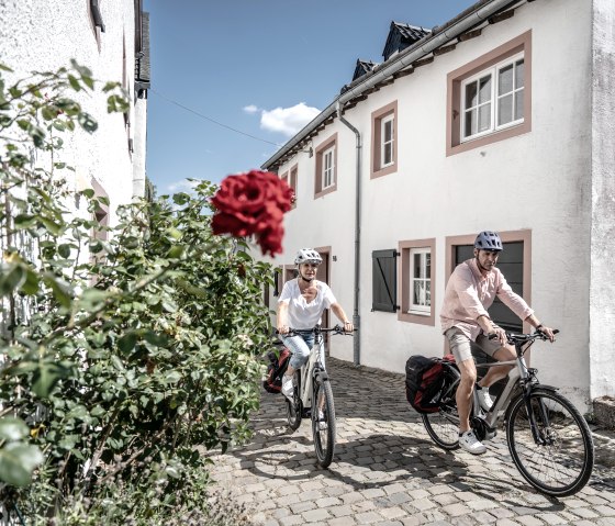 Two cyclists wearing helmets ride through the old town of the historic castle town of Kronenburg in sunny weather. In the left foreground is a rose bush, from which a red rose protrudes into the picture., &copy; Eifel Tourismus GmbH, Dennis Stratmann