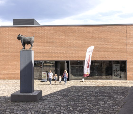 Eingang des Römerthermen Zülpich Museums mit einer Ziegenstatue auf einem Sockel. Eine Familie verlässt das Gebäude, das aus roten Ziegeln besteht., © Eifel Tourismus GmbH, Tobias Vollmer