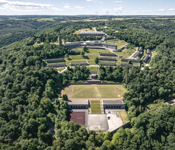 Luftaufnahme von Vogelsang IP, umgeben von gr&uuml;nen W&auml;ldern und H&uuml;geln, mit Geb&auml;uden und Wegen, die sich durch die Landschaft ziehen., &copy; Eifel Tourismus GmbH, Dennis Stratmann