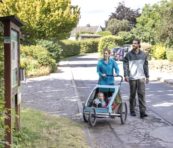 A couple pushes a baby carriage along a sidewalk in a leafy residential area. There is a sign at the side of the path., &copy; Eifel Tourismus GmbH, Tobias Vollmer