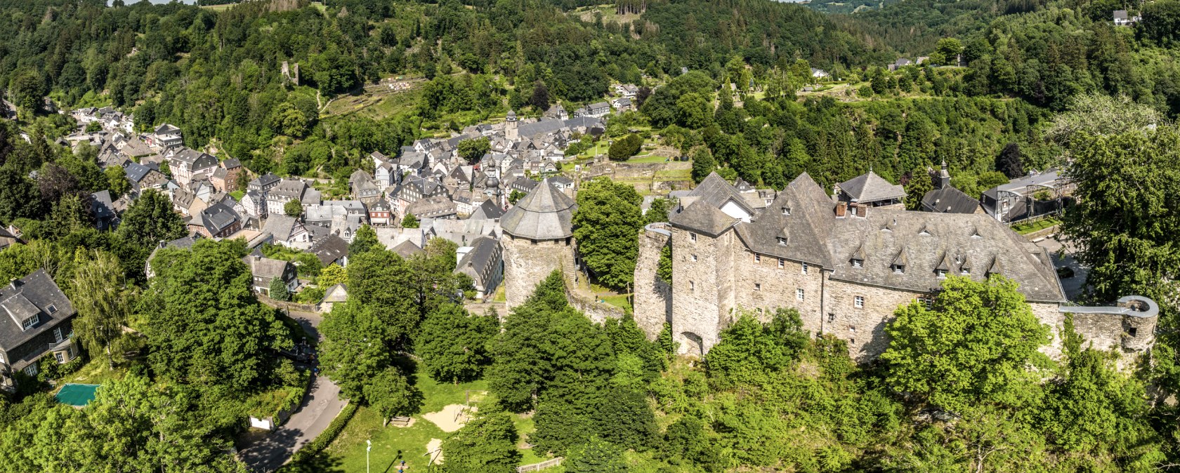 Panoramablick auf Monschau mit der Burg im Vordergrund, umgeben von gr&uuml;ner Landschaft und bewaldeten H&uuml;geln unter blauem Himmel mit Wolken., &copy; Eifel Tourismus GmbH, Dominik Ketz