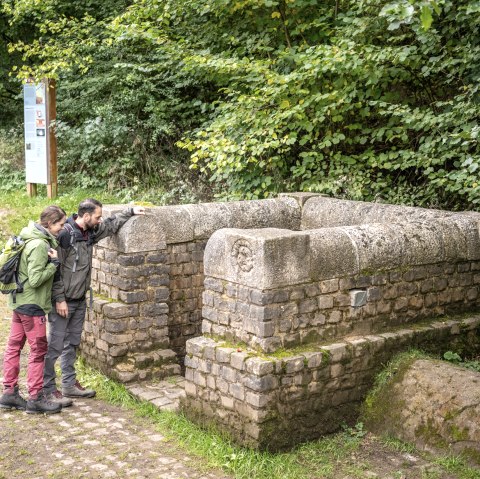 Zwei Personen betrachten einen alten Brunnen aus Stein in einem Waldgebiet. Im Hintergrund sind B&auml;ume und ein Informationsschild zu sehen., &copy; Eifel Tourismus GmbH, Dominik Ketz