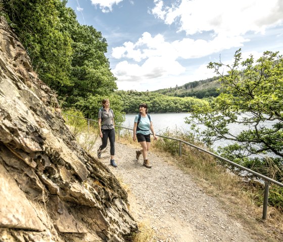 Zwei Frauen wandern auf einem schmalen Pfad entlang eines Sees, umgeben von üppigem Grün und Felsen. Der Himmel ist teils bewölkt., © Städteregion Aachen, Dominik Ketz