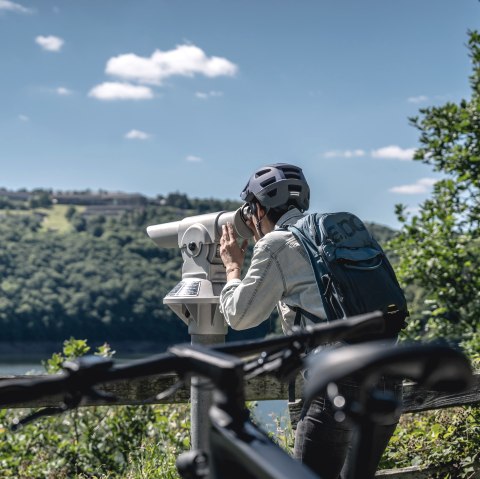 Person with helmet and rucksack looking through binoculars. Vogelsang IP can be seen in the background on a wooded hill., © Eifel Tourismus GmbH, Dennis Stratmann