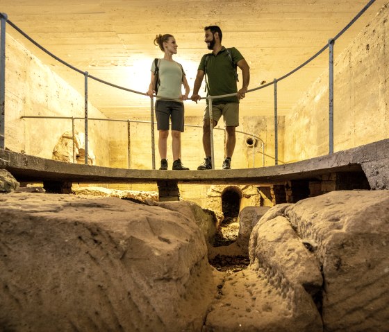 Zwei Personen stehen auf einer Plattform in einer r&ouml;mischen Brunnenstube bei Kallmuth. Die Umgebung ist aus Stein und wirkt historisch., &copy; Eifel Tourismus GmbH, Dominik Ketz