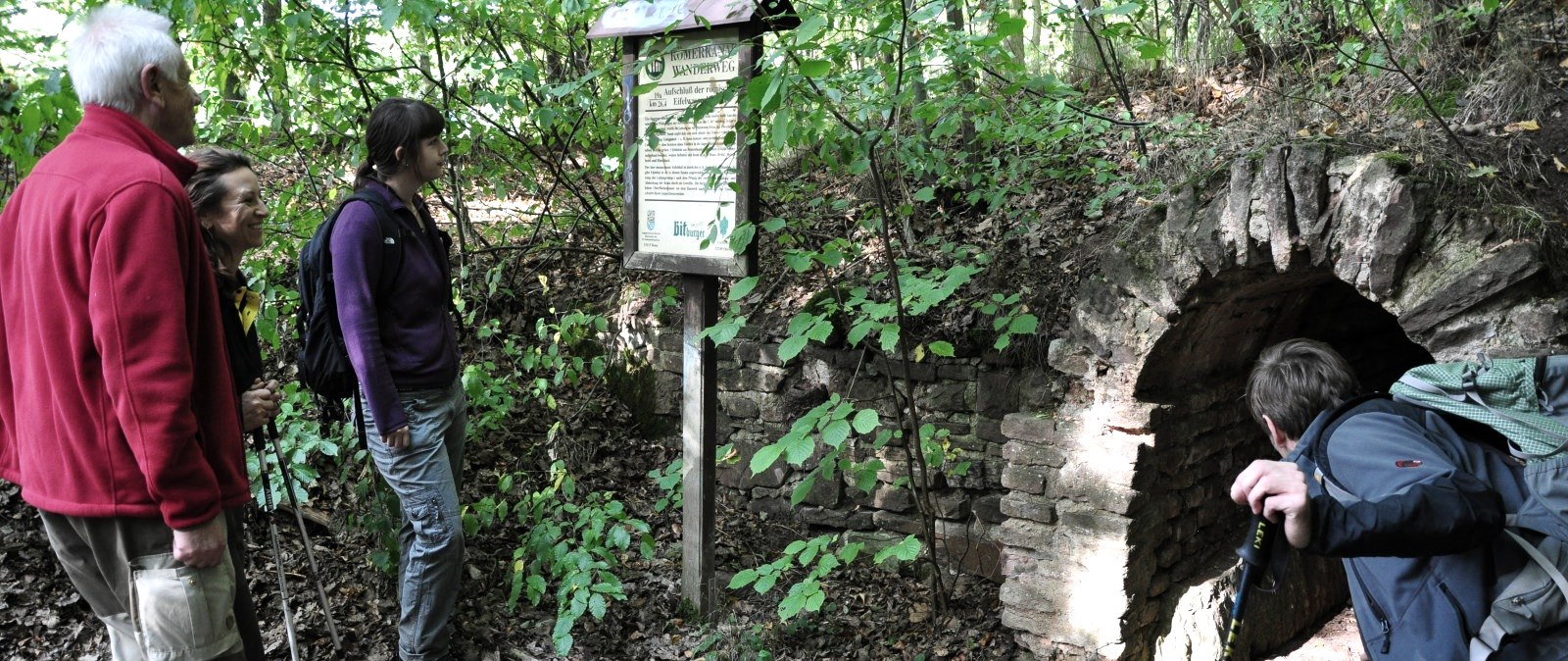 Four hikers stand in front of an information board and an old tunnel on the Roman Canal hiking trail in the forest., © Wandermagazin/ N. Glatter