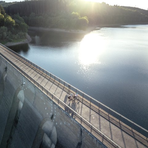 Luftaufnahme der Oleftalsperre in Hellenthal. Zwei Radfahrer fahren über die Staumauer, während die Sonne auf das Wasser scheint., © Eifel Tourismus GmbH, Dennis Stratmann