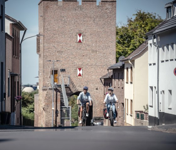 Zwei Radfahrer fahren auf einer Stra&szlig;e in Z&uuml;lpich, im Hintergrund ist das historische Bachtor zu sehen., &copy; Eifel Tourismus GmbH, Dennis Stratmann