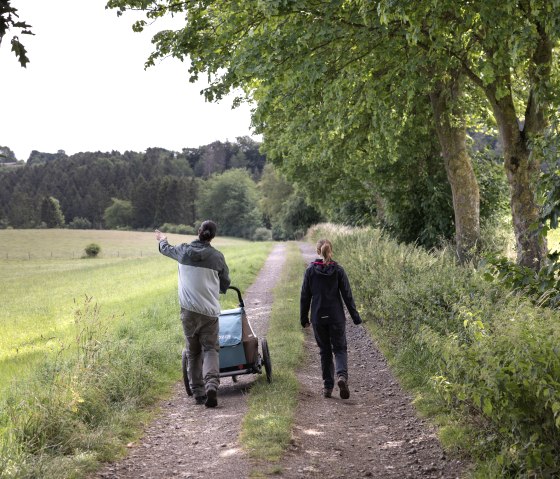 Two people are walking along a rural path surrounded by green trees and meadows. One person is pushing a cart., &copy; Eifel Tourismus GmbH, Tobias Vollmer