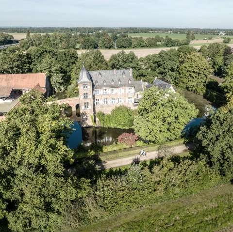 Aerial view of a castle with a moat, surrounded by trees and fields. A path leads past the castle, on which two cyclists can be seen., &copy; Eifel Tourismus GmbH, Dennis Stratmann